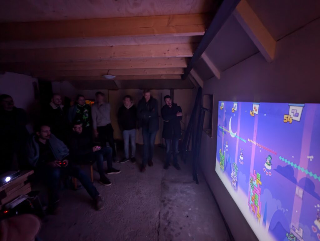 Groep mensen kijkt toe hoe een multiplayer game wordt gespeeld op een muurprojectie tijdens de Loef-LAN LAN-party.