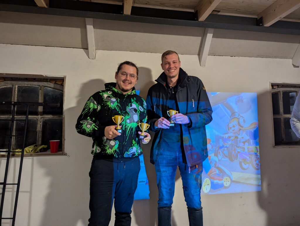 Twee winnaars poseren met gouden bekers voor een geprojecteerd beeld van Mario Kart tijdens de Loef-LAN LAN-party.