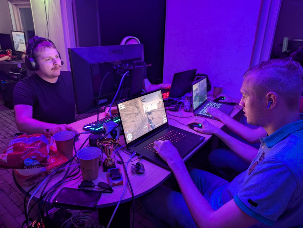 Gamers spelen intensief een shooter game tijdens Loef-LAN LAN-party, met meerdere monitoren en laptops in paarse verlichting.